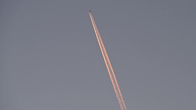 A commercial airplane high in the sky leaves long, golden sunset contrails as it flies through the evening air.