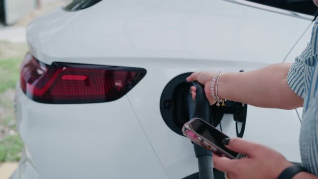 Slow motion close up of woman plugging charging cable into white electric vehicle while holding smartphone.