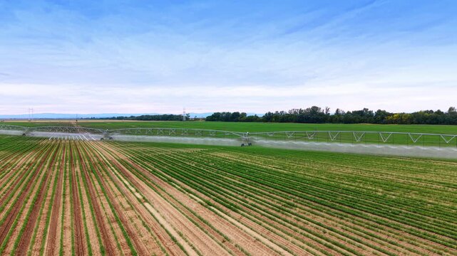 Agricultural irrigation boom extending over green field. A long metallic frame sprays water over crop rows under a cloudy blue sky.