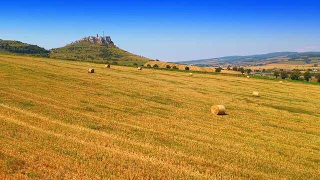Spis Castle ruin on hill above agricultural field with round straw bales. Golden harvested field with round hay bales, medieval castle ruin on hill background, clear blue sky daylight