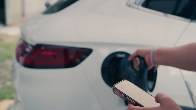 Slow motion close up of woman plugging charging cable into white electric vehicle while holding smartphone.