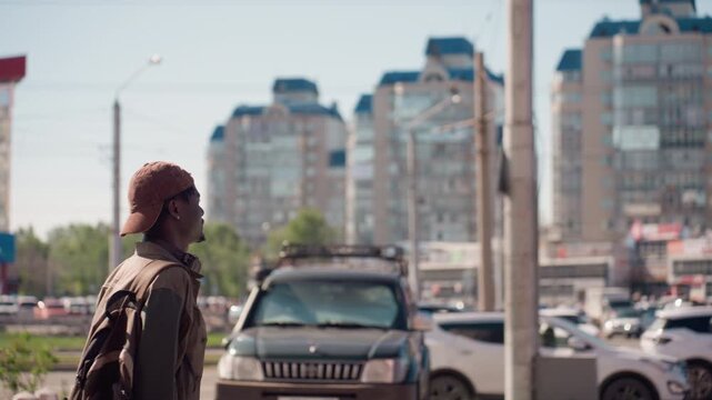 Man wearing brown cap and jacket walks along busy city street with cars passing by looking around in urban environment with tall buildings in background