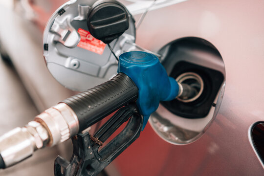 a close-up view of a person refueling a vehicle. The focus is on the individual's hand gripping a blue-handled fuel pump nozzle that is inserted into the open fuel tank of a dark car.