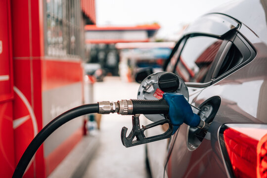 a close-up view of a person refueling a vehicle. The focus is on the individual's hand gripping a blue-handled fuel pump nozzle that is inserted into the open fuel tank of a dark car.
