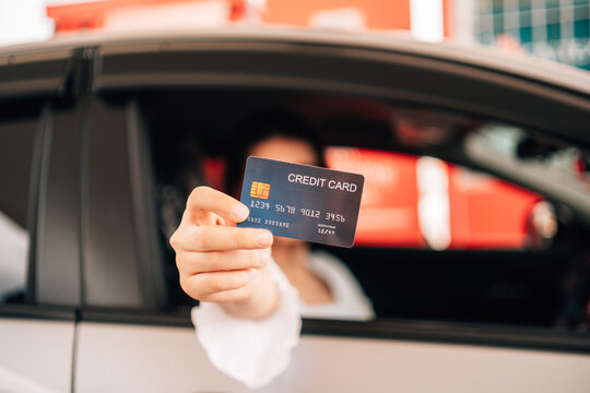 A driver's hand extends a dark credit card directly toward the camera through an open car window, with the woman in the driver's seat softly blurred in the background