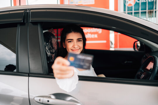 A driver's hand extends a dark credit card directly toward the camera through an open car window, with the woman in the driver's seat softly blurred in the background