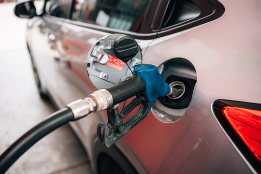 a close-up view of a person refueling a vehicle. The focus is on the individual's hand gripping a blue-handled fuel pump nozzle that is inserted into the open fuel tank of a dark car.