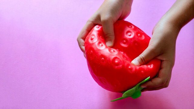 Child hands squeezing a red strawberry shaped stress toy on a pink background, finding relaxation and sensory comfort through squishy texture play for calming activity