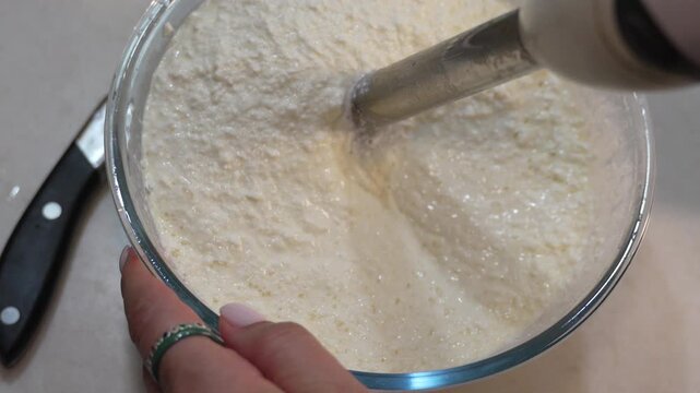 Immersion blender mixing cottage cheese in a glass bowl. Static top-down shot of kitchen food preparation.