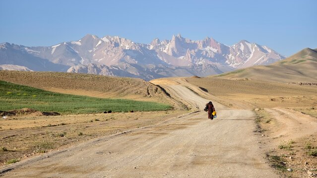 Woman in Afghanistan carry water tanks walking on dirt road toward snowcapped mountains in arid landscape