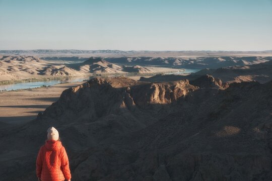 A hiker in an orange jacket looks out over a rocky canyon and the winding Ili River in Kazakhstan, ideal for active lifestyle branding, adventure travel promotions, and ecotourism layouts.