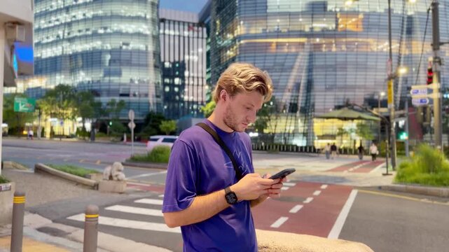 Young Blond Man Standing at Pedestrian Crossing in Seoul at Night Using Smartphone &ndash; Urban Street Scene with City Lights