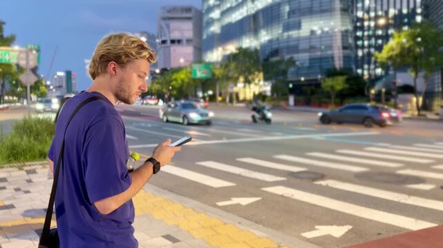 Young Blond Man Standing at Pedestrian Crossing in Seoul at Night Using Smartphone &ndash; Urban Street Scene with City Lights