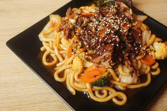 Asian style beef noodles with vegetables and sesame seeds on black plate. Udon pasta broccoli carrots and cauliflower form savory meal background. Restaurant lunch dish with rich brown sauce.