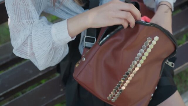 Close up of young woman sitting on outdoor bench unzipping her brown leather backpack with gold hexagonal embellishments to reveal and remove crocheted items. Woman wears white lace blouse