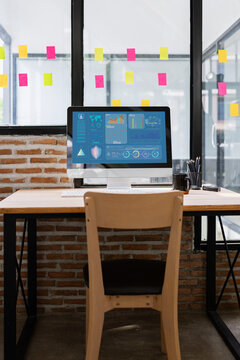 A modern office workstation with a computer displaying data analytics and charts on the screen, set up on a wooden desk with a brick wall and sticky notes in the background.