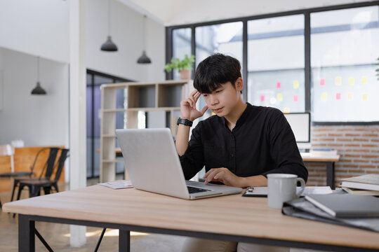 Stressed young Asian businessman working on a laptop in the office, feeling exhausted, overwhelmed, and experiencing a headache from heavy workload.