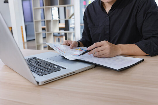 A young Asian businessman working at a desk in a modern office, writing notes on a clipboard while using a laptop.