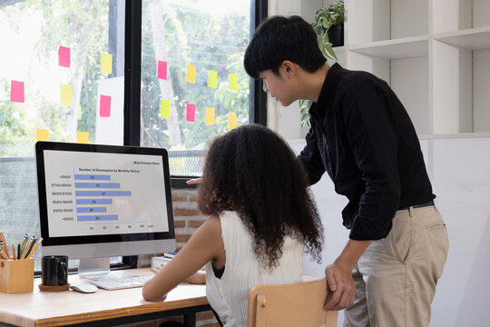 Asian male colleague standing and discussing business data charts and statistics on a computer monitor with his multi-ethnic female coworker in a modern office.