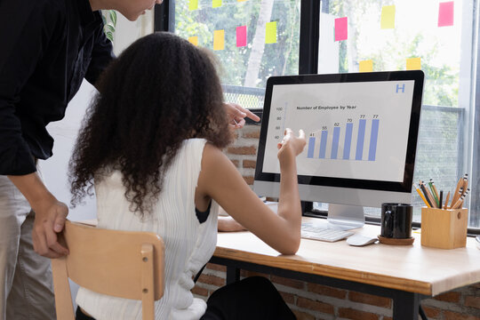 Asian male colleague standing and discussing business data charts and statistics on a computer monitor with his multi-ethnic female coworker in a modern office.