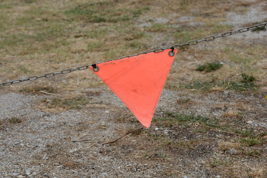 An orange triangular warning flag hangs from a metal chain fence stretched across a gravel and grass ground.