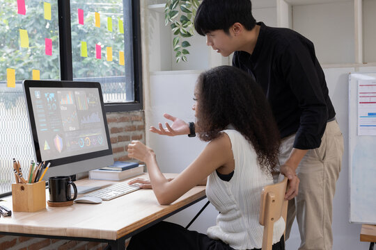 Asian male colleague standing and discussing business data charts and statistics on a computer monitor with his multi-ethnic female coworker in a modern office.
