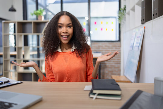 A cheerful young businesswoman with curly hair sitting at an office desk, gesturing and welcoming clients or presenting work in a modern workplace.