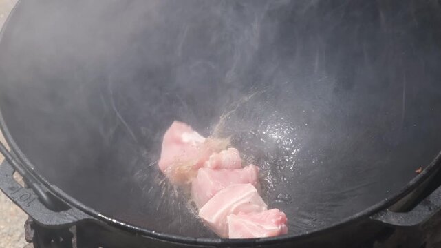 Raw pork meat frying in hot oil inside a traditional cast iron cauldron outdoors.