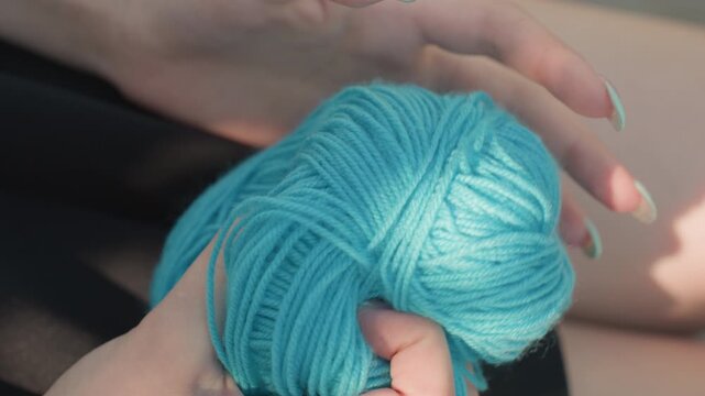Close up of hands holding and manipulating ball of turquoise yarn showcasing texture detail as fingers gently grasp and unwind material. Soft lighting highlights fiber strands