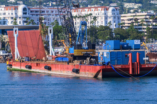 Cannes, France - July 30, 2026: A large red and blue construction barge with a crane and equipment floats on blue water near a city