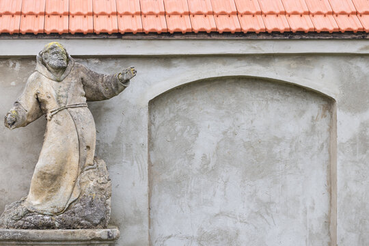 Ancient weathered stone statue of a monk or saint near wall with arched niche and tiled roof