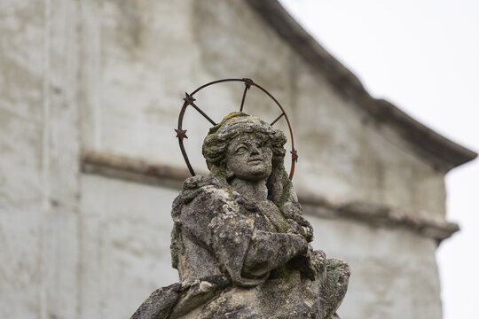 Ancient weathered stone statue of Virgin Mary with metal halo and stars against light wall background