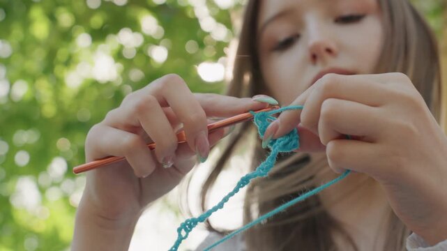 Close up of young woman crocheting with blue yarn and orange crochet hook in soft natural light. Her hands manipulate material showing detailed finger movements as woman creates textile