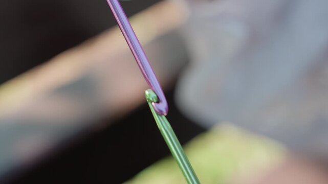Close up of hand holding colorful crochet hooks looping yarn in soft lighting. focus remains on detailed interaction between metal tools and fabric material. Background is intentionally