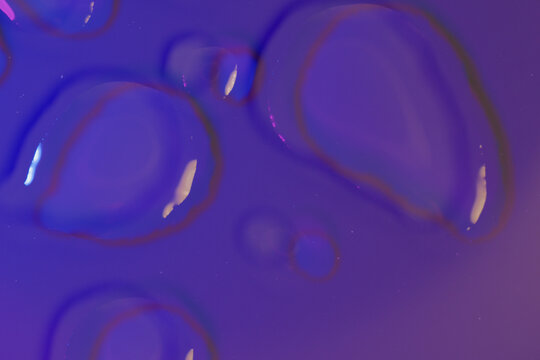 Macro shot of elongated water droplets with bright reflections on dark violet purple background. Futuristic wet texture for night repair skincare, hyaluronic serum, luxury cosmetics, biotech concepts.