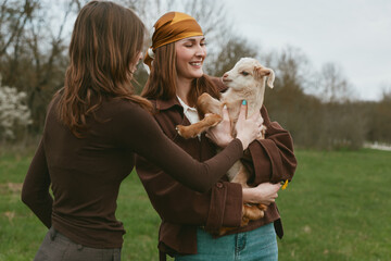 Naklejka premium Two women outdoors interact with a small goat kid, smiling as they cradle the young animal. Natural field setting emphasizes lifestyle, nature, and gentle companionship in a warm, candid moment.