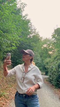 Active woman wearing a cap walking on a countryside path through a lush green forest, looking at her smartphone for navigation or taking selfies during her outdoor adventure and trekking