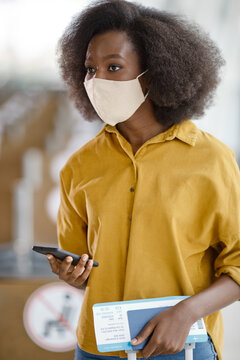 A young Black woman with an afro hairstyle wears a beige face mask and a yellow shirt while holding a smartphone and a boarding pass at an airport terminal
