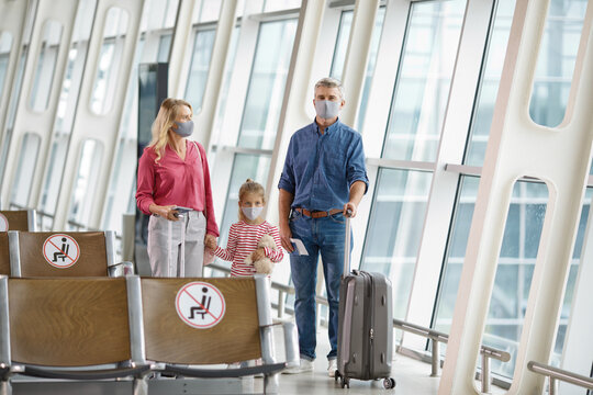 A family with a child wearing face masks and carrying luggage walks through a modern airport terminal, adhering to safety protocols