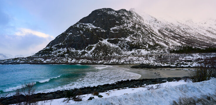 Beautiful scenic viewpoint of a coastal highway winding next to the blue sea in Lofoten, Norway. Majestic winter landscape with grand snow-covered mountains under a dramatic overcast sky in Arctic Sca