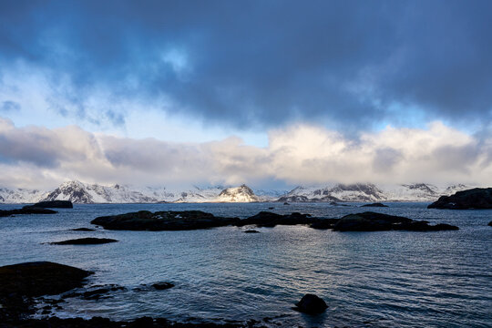 Beautiful scenic view of a traditional Norwegian fishing village with red wooden cabins by the fjord in Lofoten, Norway. Majestic snow-covered mountains under a dramatic winter sky in Arctic Scandinav