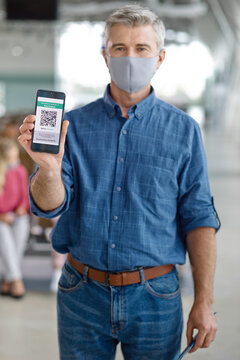 A middle-aged man in a blue checkered shirt and jeans displays his digital COVID-19 vaccination record on his mobile phone while wearing a grey face covering