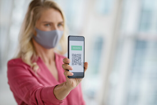 A woman wearing a face mask displays her mobile phone screen showing a digital vaccination record with a QR code for verification
