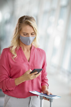 A woman in a pink shirt and grey mask looks at her smartphone while holding travel documents and a suitcase handle