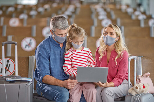 A family of three wearing protective face masks sits together in an airport waiting area, looking at a laptop computer