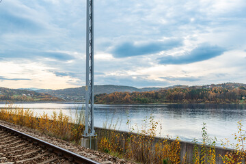 Electrified railway track runs beside Jezioro Mucharskie reservoir with forested Beskid hills and autumn foliage across the water. Scenic transport infrastructure image combining railroad engineering, © Volha