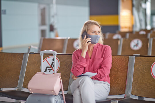 A woman with blonde hair sits in an airport waiting area, wearing a protective face mask and speaking on her mobile phone with luggage beside her