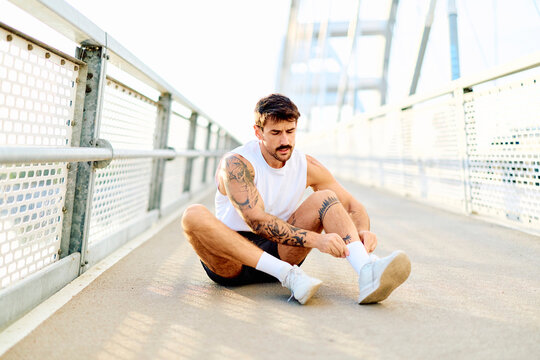 Young athletic man with tattoos resting on a bridge, adjusting socks and preparing for or recovering from a run or exercise