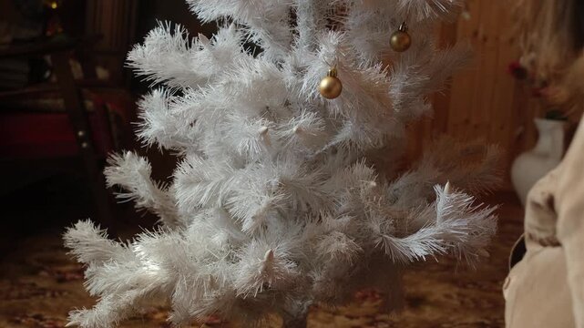 Woman decorating white Christmas tree with Christmas balls in cozy home.