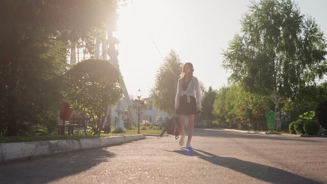 Young woman walks down sunlit street carrying backpack wearing casual clothing with headphones. scene features outdoor elements like trees greenery and distant structures under bright